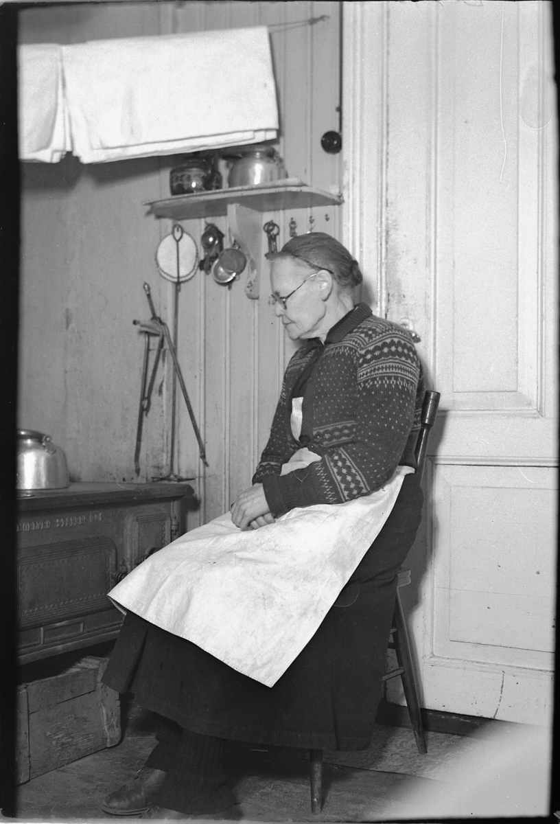 Håkon Prestkværn, Interiør, kjøkken. Ukjent dame sitter ved ovn med kaffekjele. Ovn produsert av Moelven støperi.