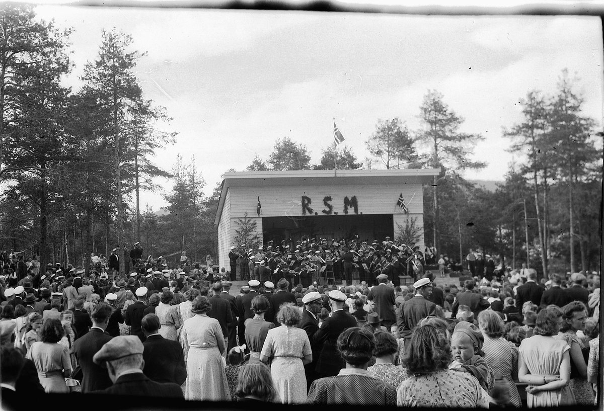 Håkon Prestkværn, Ringsaker sang- og musikklagsstevne, Moelv 1948. Friluftskonserten i Folkets Hus park. Stor gruppe mennesker hører musikkorps spille Folkets Hus i Moelv.