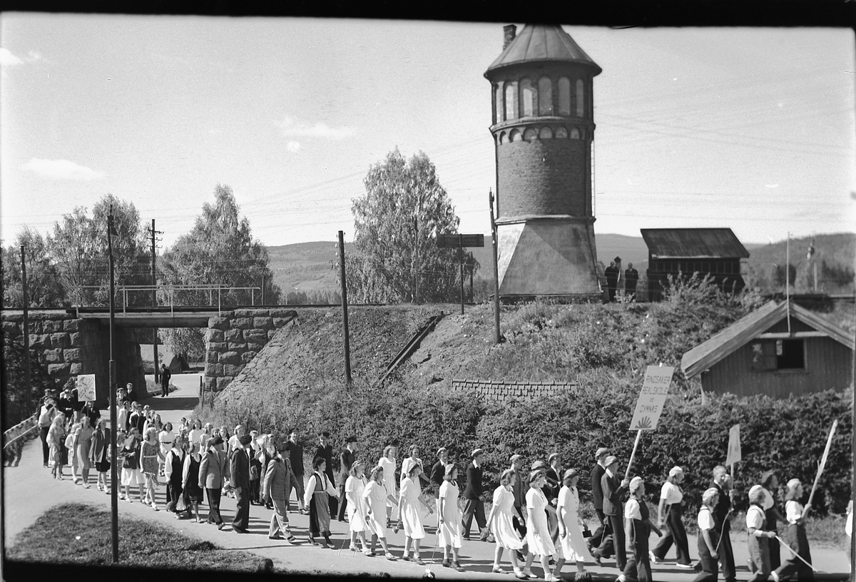 Håkon Prestkværn, 17. mai-tog i Storgata, Moelv. Vanntårnet ved Moelv jernbanestasjon i bakgrunnen.