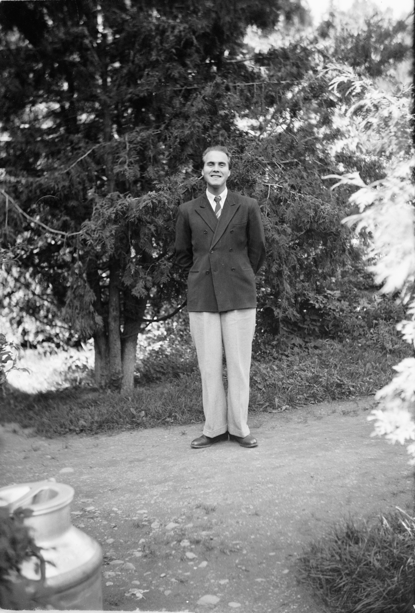 Håkon Prestkværn, Mann i dress ved et grantre med et melkespann i forgrunnen.