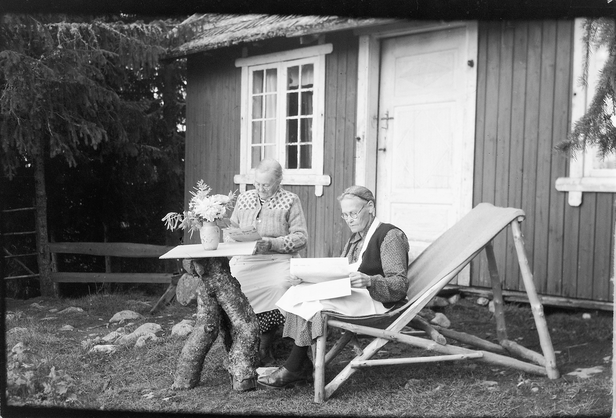 Håkon Prestkværn, To eldre kvinner utenfor ei hytte. f. v. ukjent og Gunda Prestkværn, hagemøbler, fluktstol, bord, avislesing,