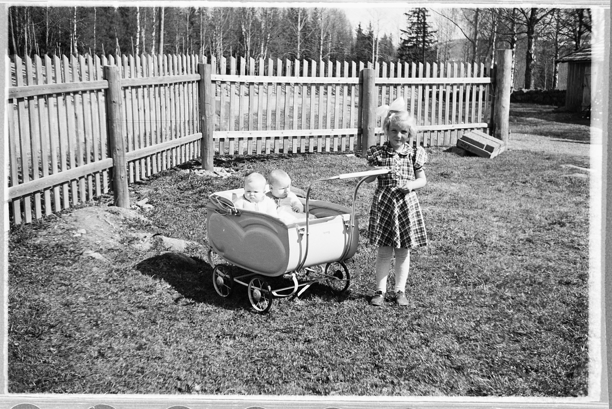 Håkon Prestkværn, To babyer i en bagnevogn og ei jente med sløyfe i håret. Tvillingene i vogna er Knut og Bjørg Svenskerud f. 1949. Jenta med sløyfe er Inger Svenskerud f. 1945. Bildet er tatt ved Åsmarka kirkestue.