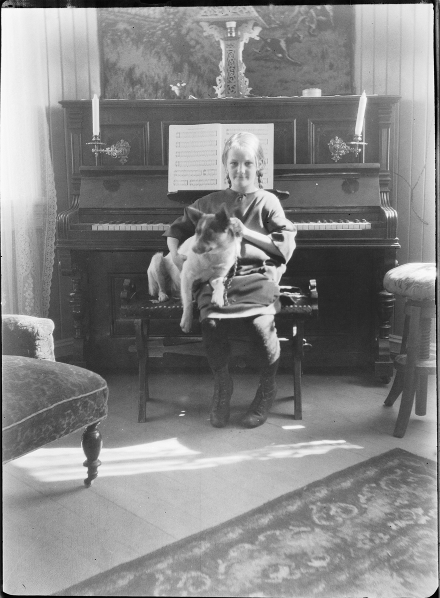 Ole M Martinsen, Interiør, stue. Jente med en hund på fanget sitter foran et piano.