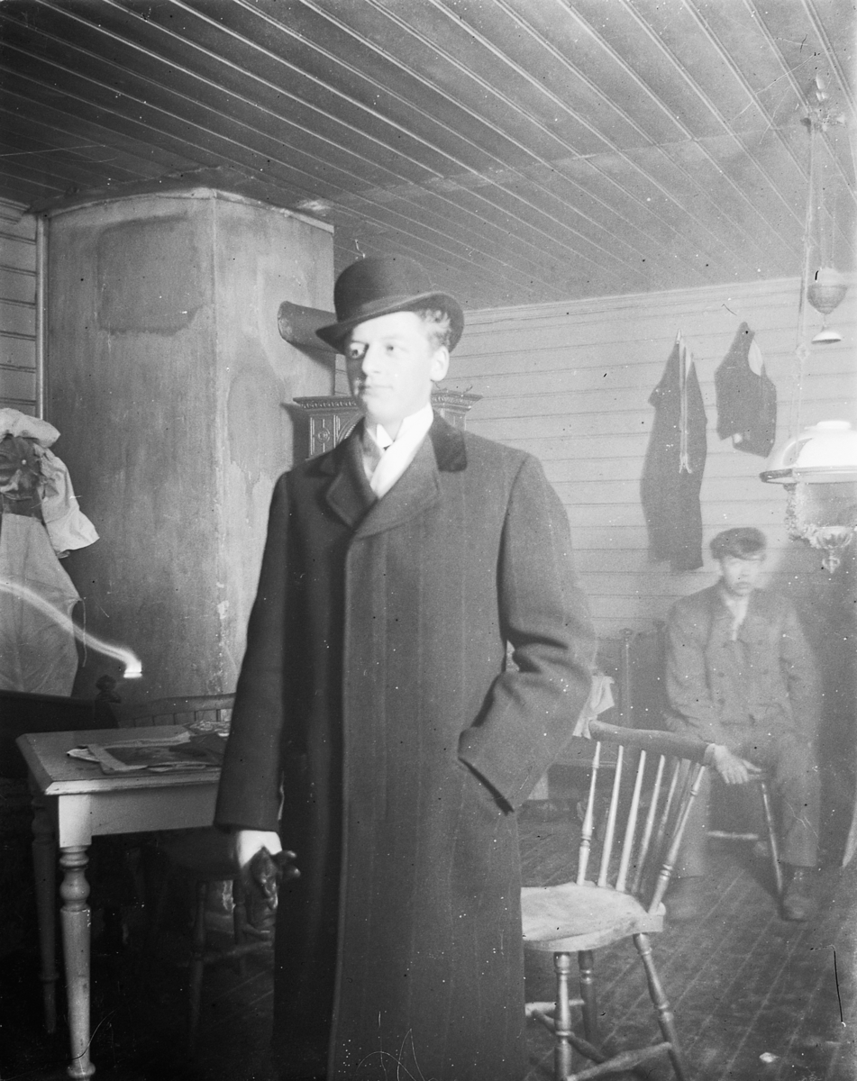 Johs. (Johannes) Johannessen, Interiør, hytte. Ukjent mann med skalk og frakk.
