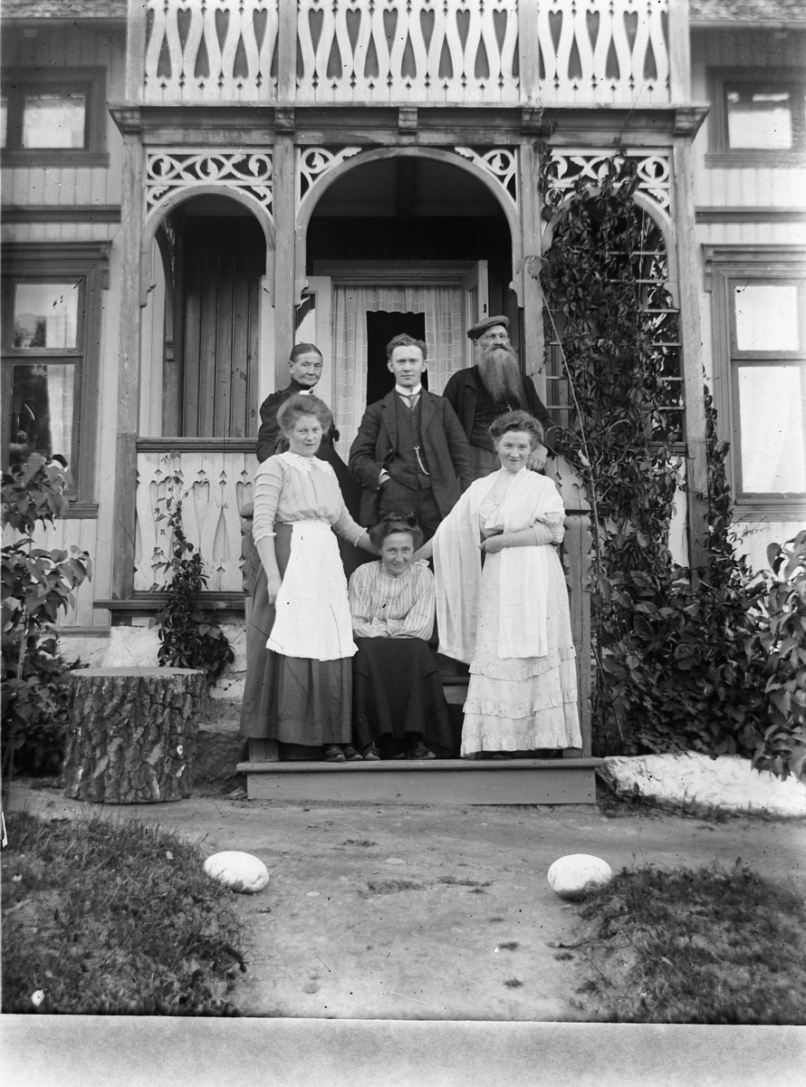 Ragnvald Andersen, Gr. 6. Familien Danielsen. To menn og fire kvinner foran veranda. Sveitserhus med hage. Kjøpmann Danielsen, Lundberg, Haraldstad, Furnesvegen. Fotografens svigerfamilie, han sjøl i midten. Fru Pernille Danielsen, F. S. Fokstuen, kjøpmann Danielsen, Olga Andersen og Dagmar Danielsen, døtre av kjøpma…