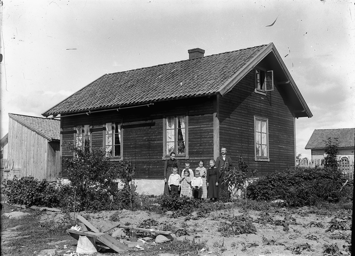 Ragnvald Andersen, Gr. 8. Familie i hagen foran hus. I bakgrunnen P. G. kolonial, manufaktur, glass.