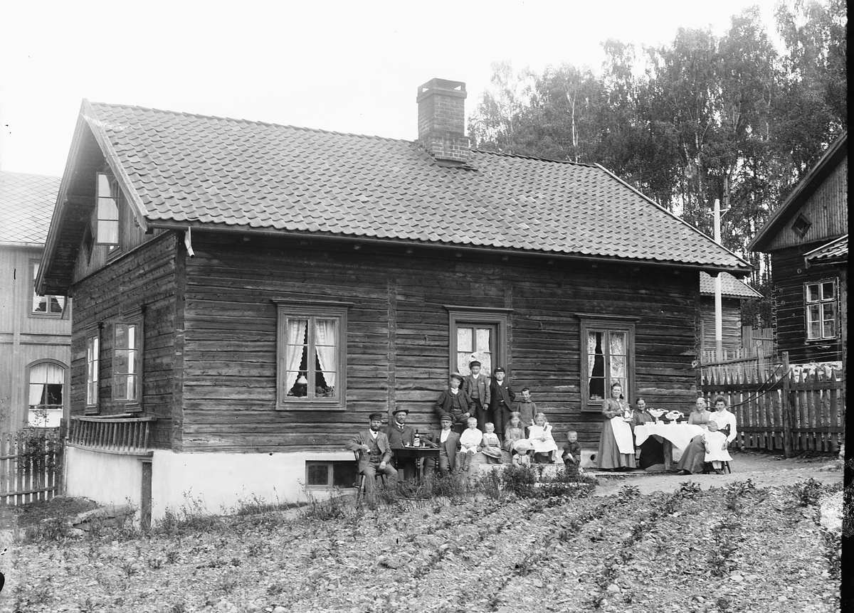 Ragnvald Andersen, Stor gruppe menn, kvinner og barn sitter i husveggen og drikker kaffe.
