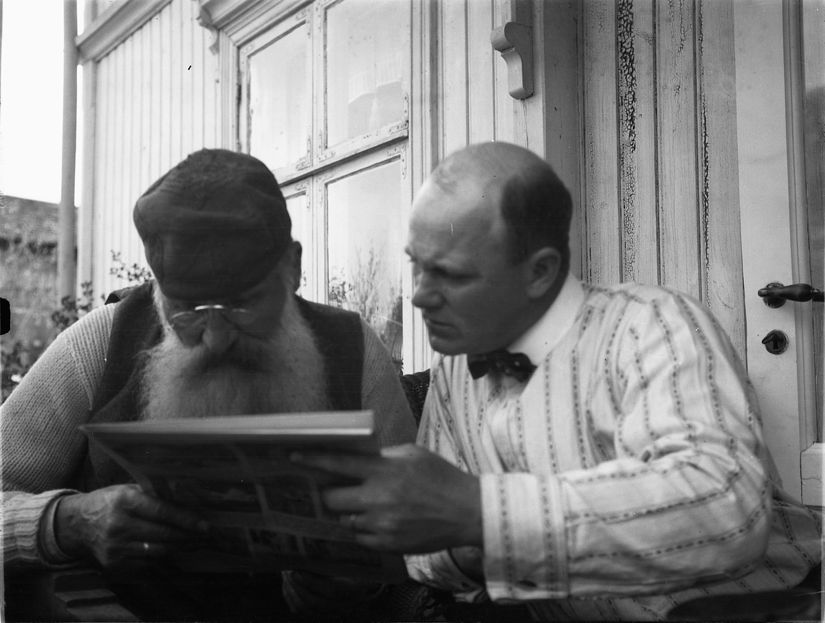 Ragnvald Andersen, To menn leser i ei avis ved inngangsdøra til et hus. Tv: Oluf Danielsen, Lundberg, Furnesvegen.