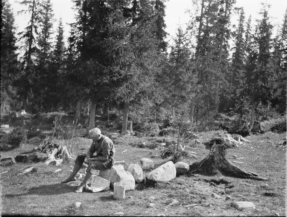 Ragnvald Andersen, Ukjent mann sitter på en stein i skogen. Skreller poteter?