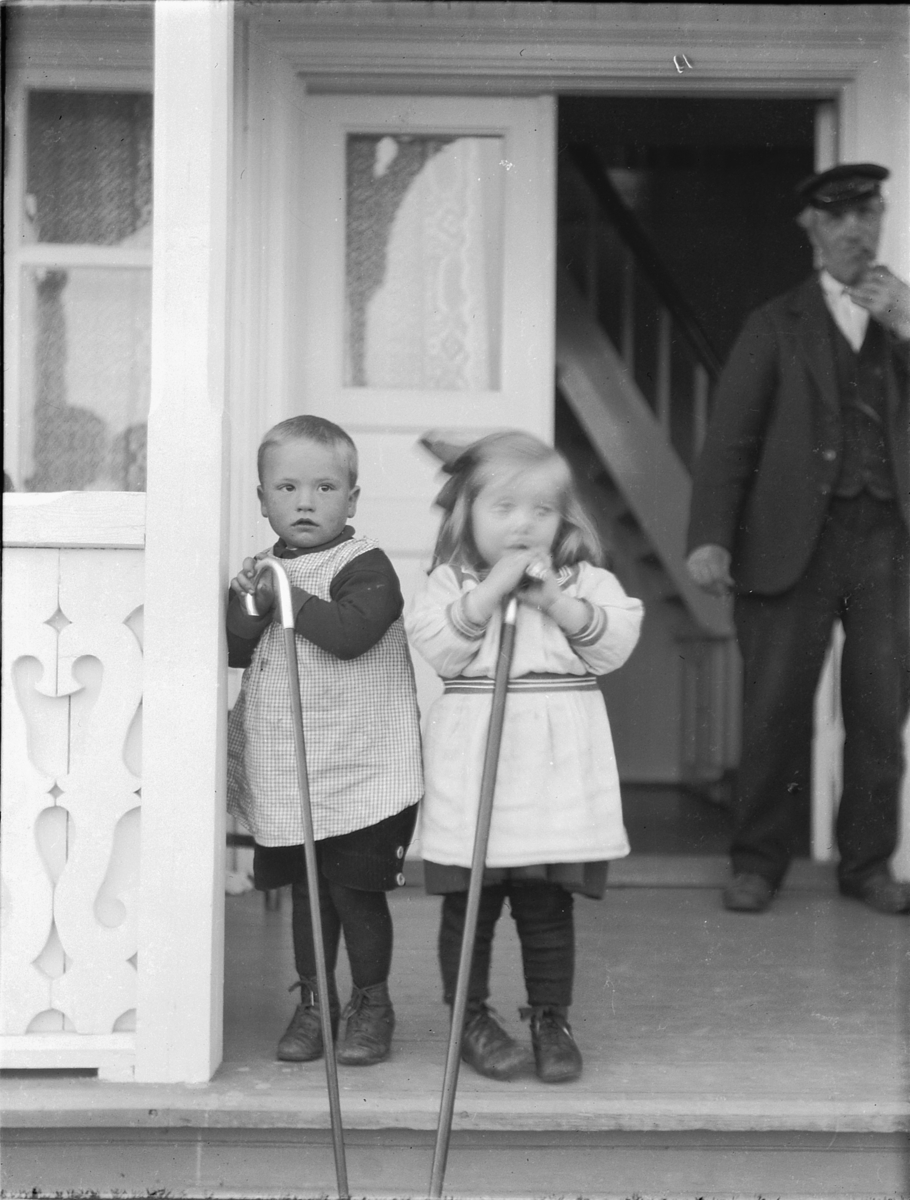 Ragnvald Andersen, To barn med hver sin stokk ved ei inngangsdør. Ukjent eldre mann i bakgrunnen.