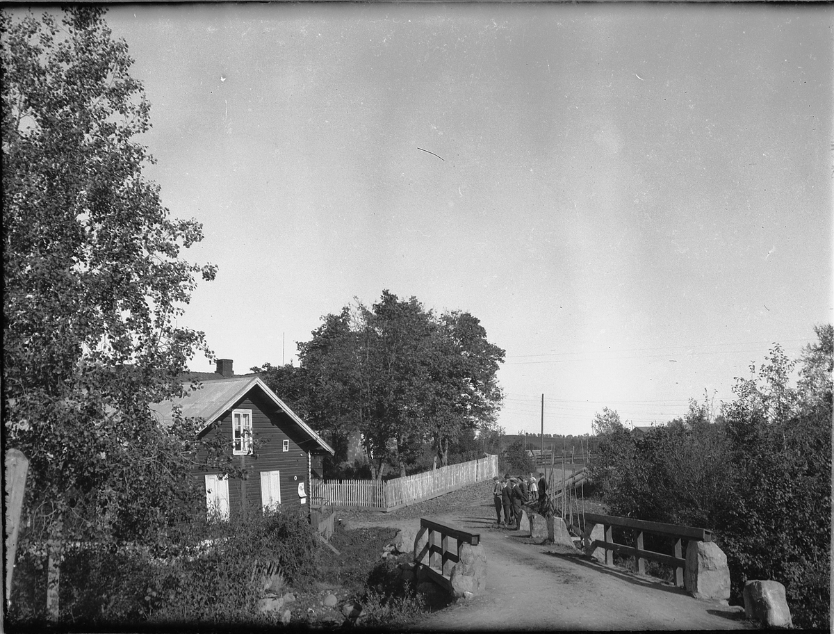 Ragnvald Andersen, Gruppe mennesker på en vei, ved ei bru og et hus.