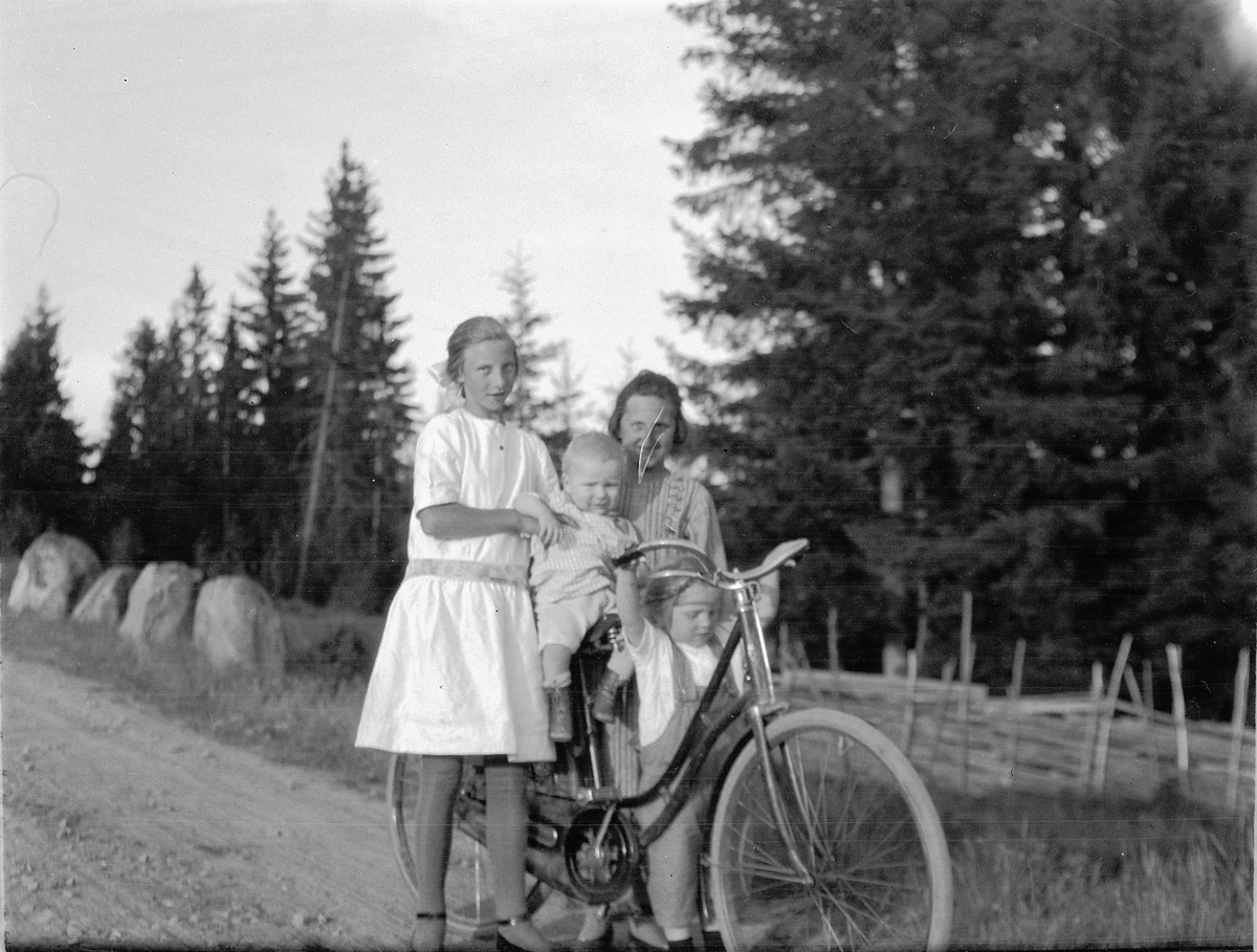Ragnvald Andersen, To jenter og et barn ved en sykkel på en landevei i skogen.