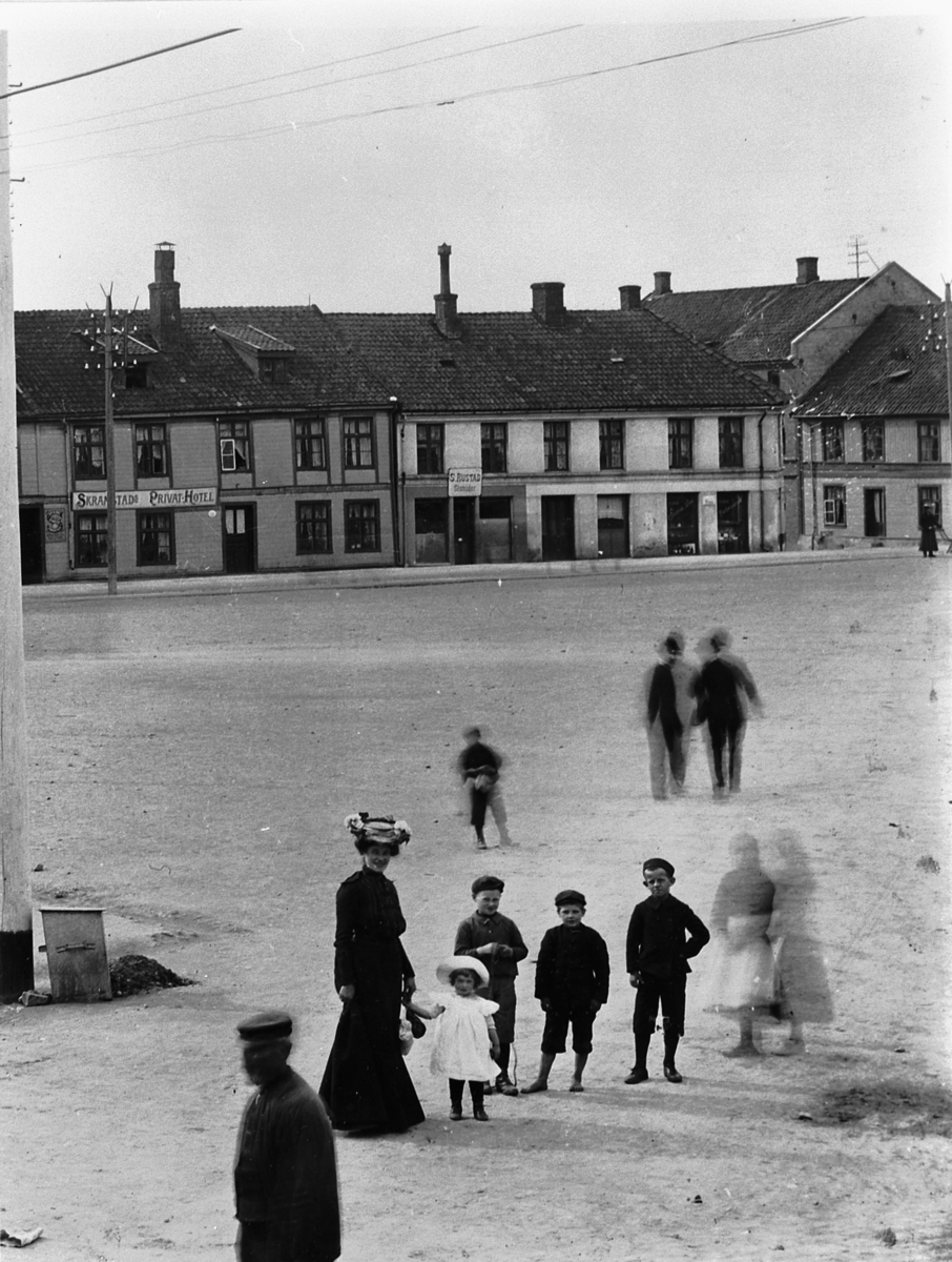 Hans Emil Erichsen, Østre Torg i Hamar. Bebyggelse langs Torggata. Skramstad Privathotell, Skomaker S. Rustad. A. Hage Colonial, Vine og fødevarer. Voksne og barn på torget. Utsnitt.