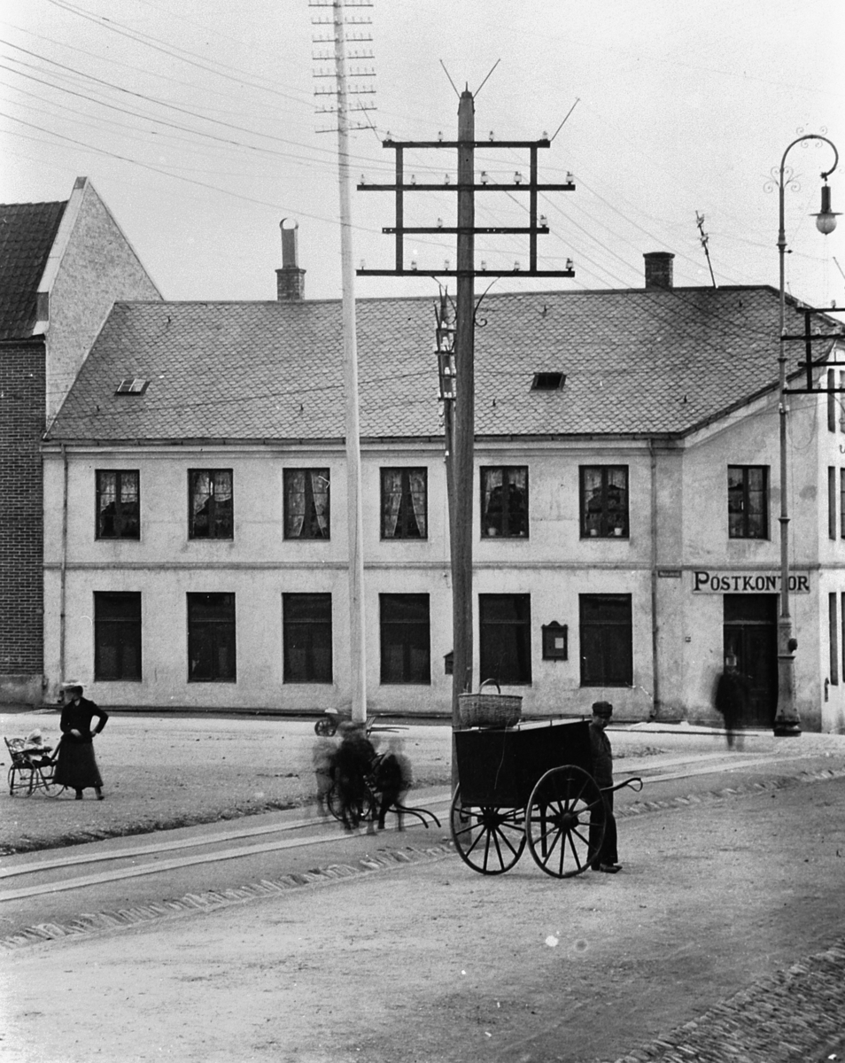 Hans Emil Erichsen, Østre Torg i Hamar med bebyggelsen langs Vangsvegen. Hamar Postkontor. Mann med håndkjerre. Lysstolper og gatebelysning. Utsnitt.