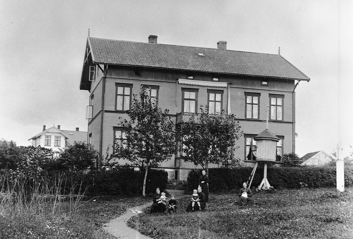 Hans Emil Erichsen, Parkgata i Hamar. Huset til stasjonsmester Christian Fredrik Lund, bygget i 1878 før Parkgata ble opparbeidet. Da gata ble laget lå huset i veien så det måtte flyttes 90 grader. Huset ble revet i 1968. Gruppe barn og voksne ute i Hagen. Dueslag til høyre. Til venstre i bakgrunnen Parkgata 42, bygge…