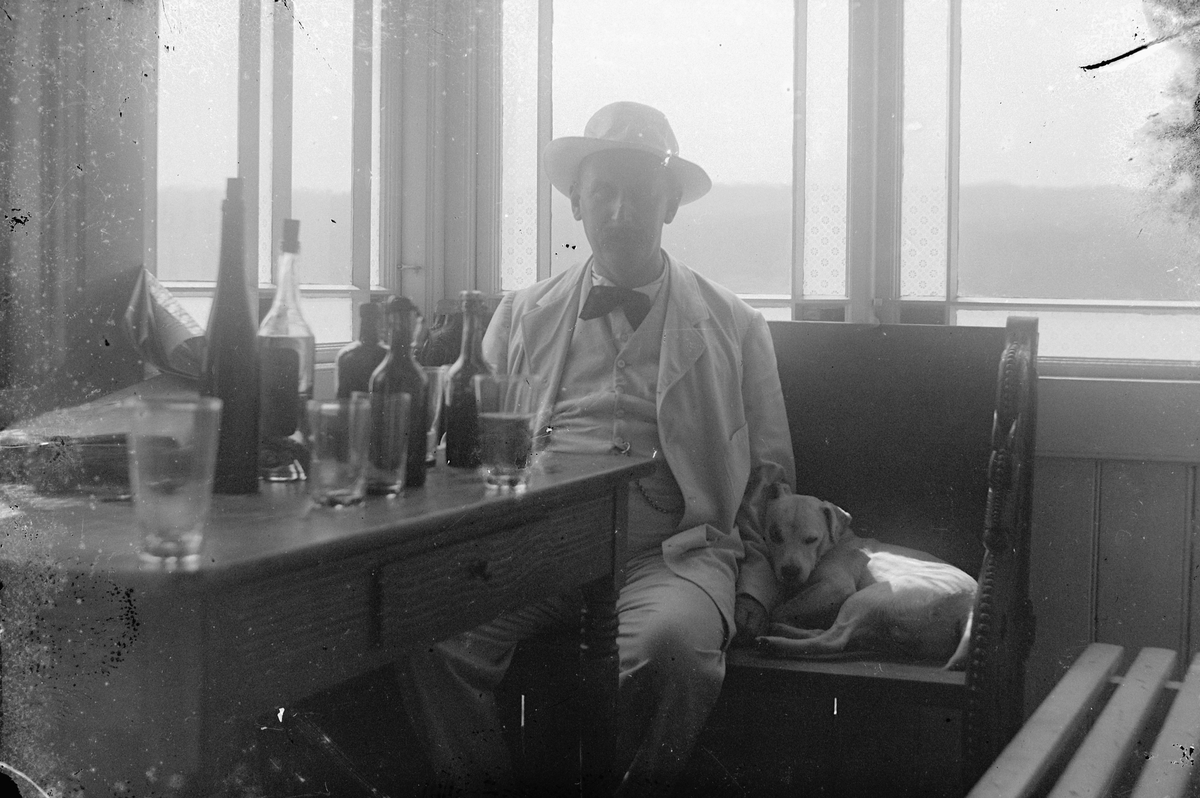 Hans Emil Erichsen, Ukjent mann med hund i glassveranda. Flasker og glass på bord.