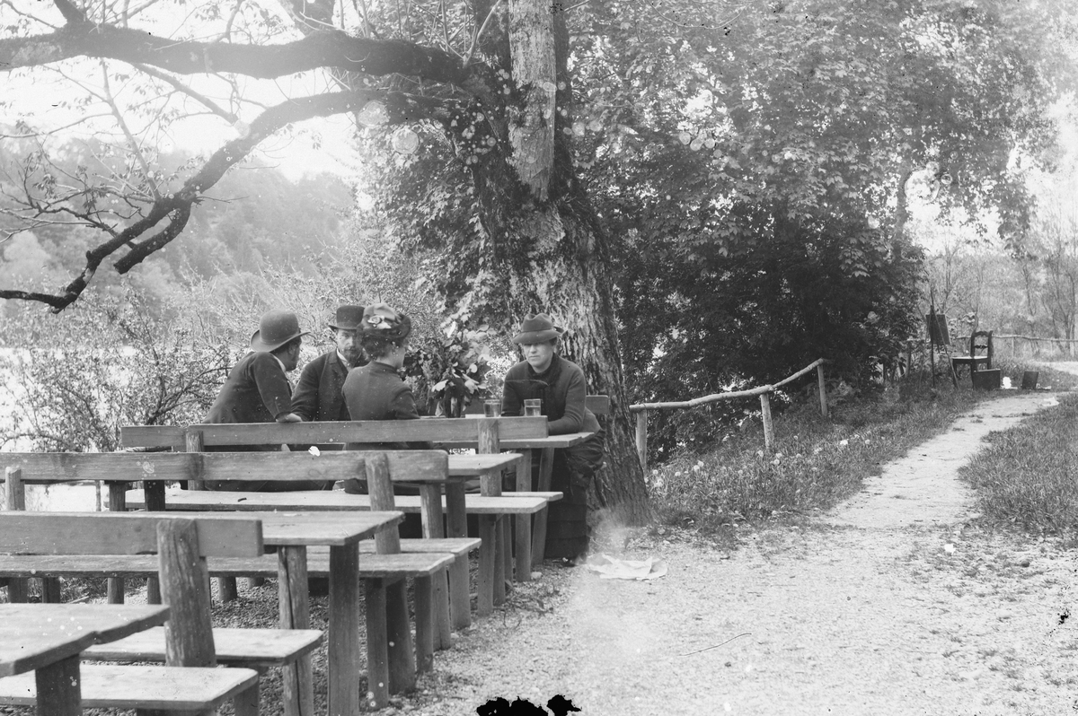 Hans Emil Erichsen, Gruppe voksne ved benker i park. Staffeli og malersaker i bakgrunn.