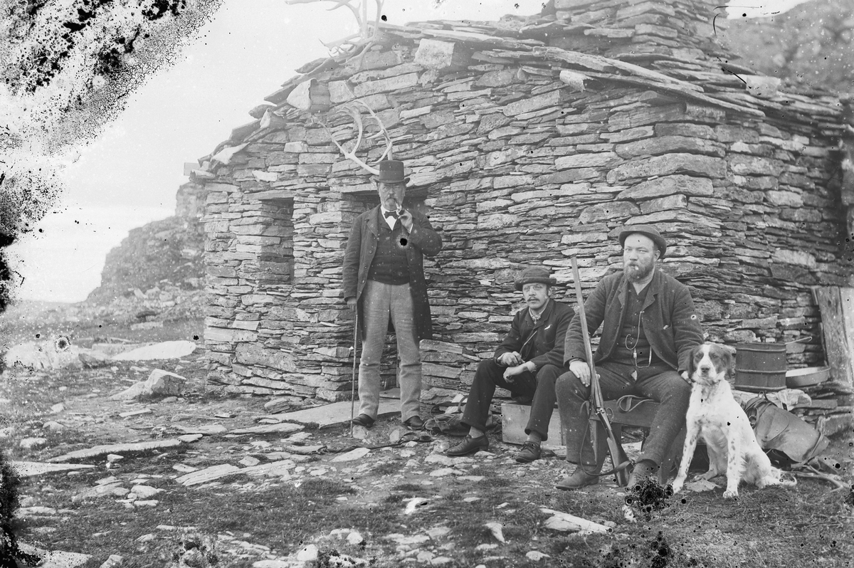 Hans Emil Erichsen, 3 menn med hund og gevær ved Friisbua på Ringebufjellet. Fra venstre er J.A. Friis, Ludvig Skramstad, ukjent. Denne steinhytta ble bygget av forfatteren og professoren J. A. Friis i 1878. Friis ferierte her i fjellet i mange år, noe han har beskrevet i boka ”Til fjeld i Ferierne. Jæger og Fiskerliv…