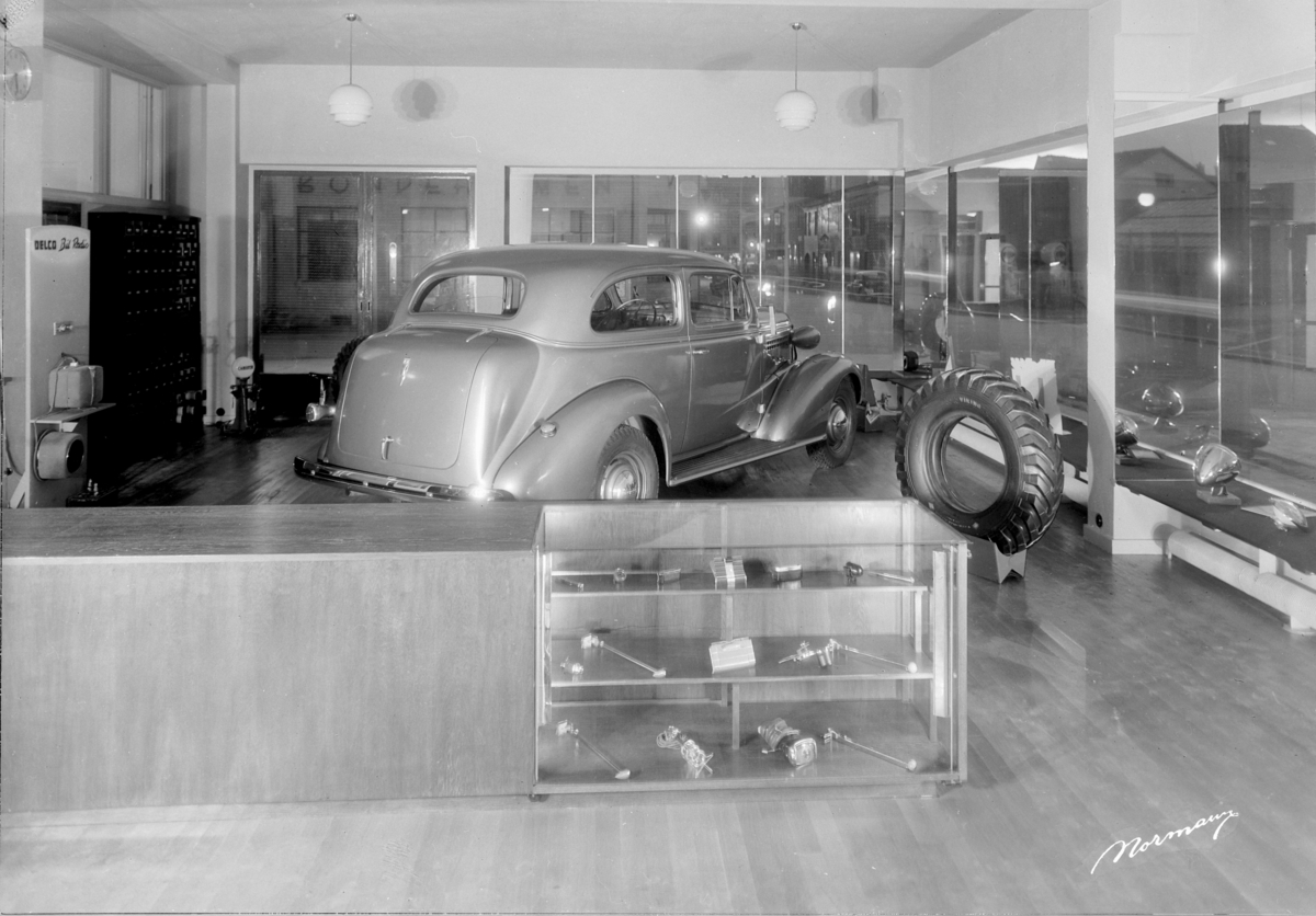 Normann Fotoatelier, Oustad Mek. Verksted A/S. Hamar. Interiør, Utstillingslokale. Bilen er en Chevrolet 1937-38. Disse to årsmodellene karakteriseres av den rette, skrånende profilen nederst på pansersiden med fortsettelse et stykke inn på døra. Bilbutikk.