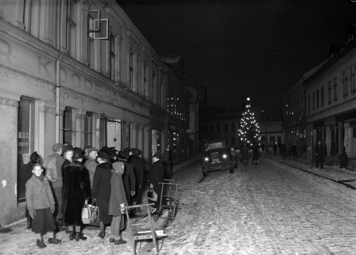 Normann Fotoatelier, Matkø under krigen. 23. 12. 1943. Strandgt. / Torggt. 52 utenfor Hamar Kooperative i Hamar. Kl. 7 - 7. 30. I bakgrunnen skimtes juletreet ved Triangelparken. Lastebil med blendede frontlykter i bakgrunnen. Spark.