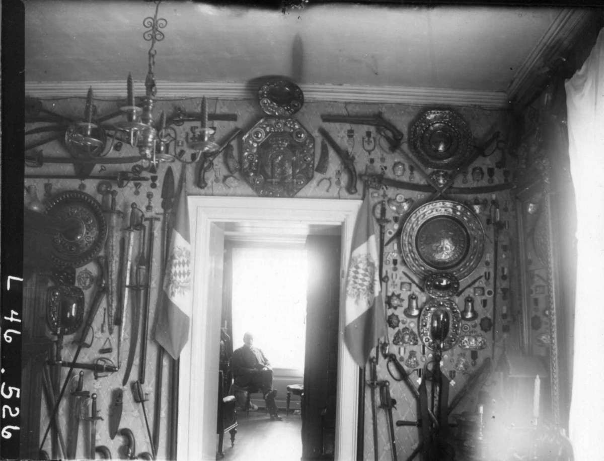 Fredrik Emil Faye, Interiør, Dal gård, Ullensaker. Vegg i gjennomgangsrom med opphengte våpen, skjold, taklampe. Kammerherre Faye sitter i rom innenfor. Ca. 1898. Fra serie fotografert av kammerherre Fredrik Emil Faye (1844-1903), gårdens eier.