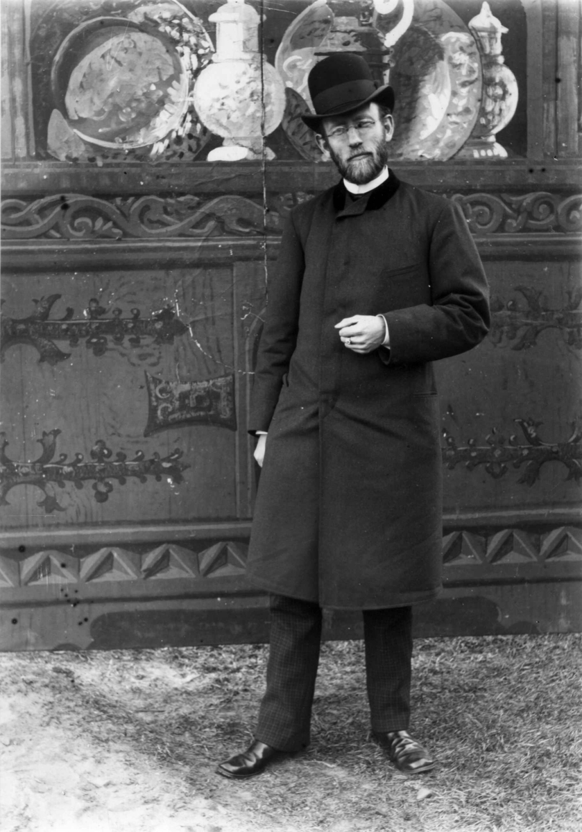 Fredrik Emil Faye, Portrett, Henrik August Grosch (1848-1929), direktør for Kunstindustrimuseet, Oslo, stående utendørs foran malt vegg eller bakteppe med motiv skap med oppstilt keramikk. Fra portrettserie av personer som bodde på eller besøkte Dal gård, Ullensaker, fotografert av gårdens eier, kammerherre Fred. Emi…