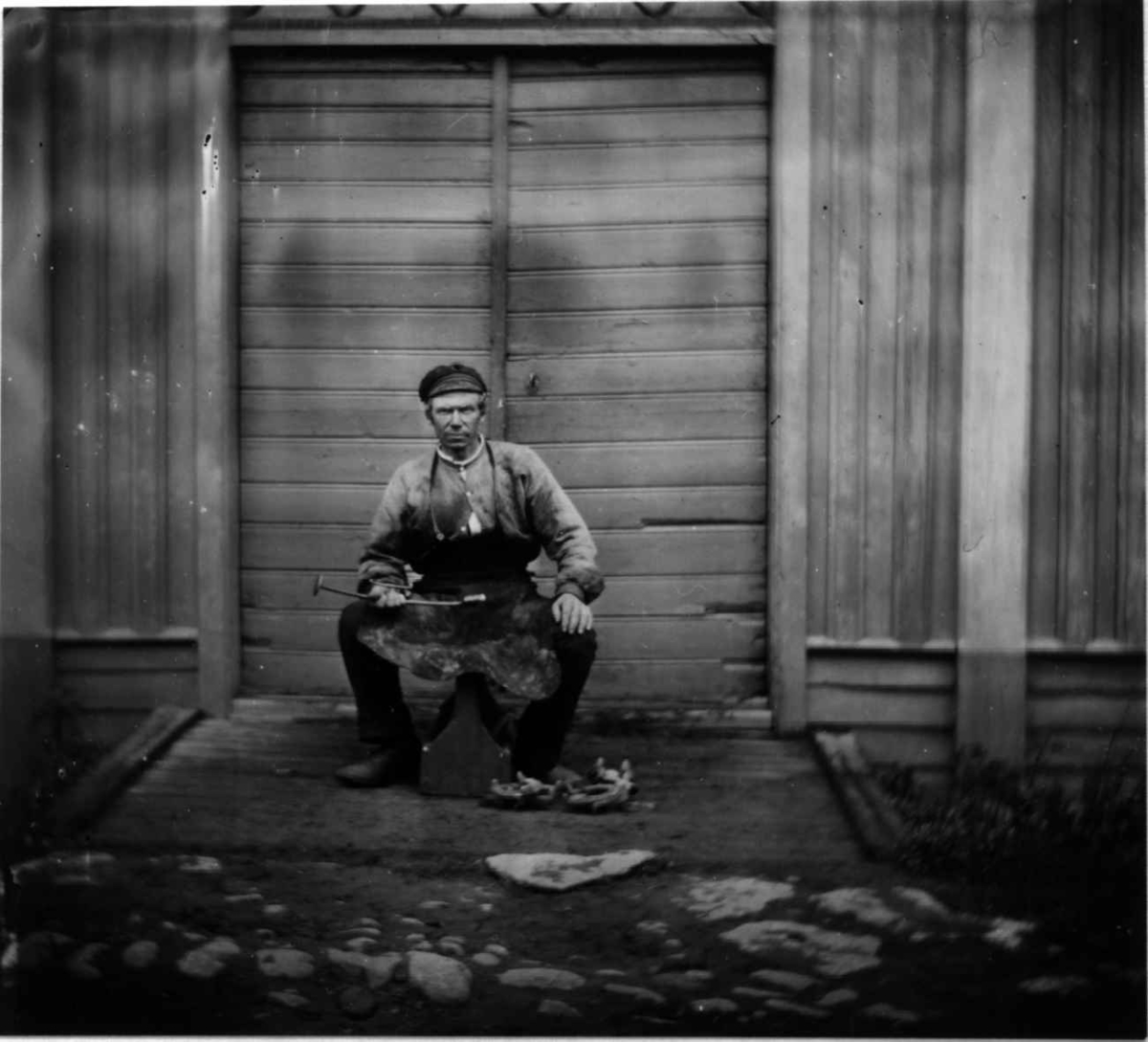 Fredrik Emil Faye, Hesteskosmed, Dal gård, Ullensaker. Mann i arbeidstøy med arbeidsredskaper poserer foran inngangsparti, ca. 1880. Fra portrettserie av personer som bodde på eller besøkte Dal gård, Ullensaker, fotografert av gårdens eier, kammerherre Fredrik Emil Faye (1844-1903) i årene 1875-1900.
