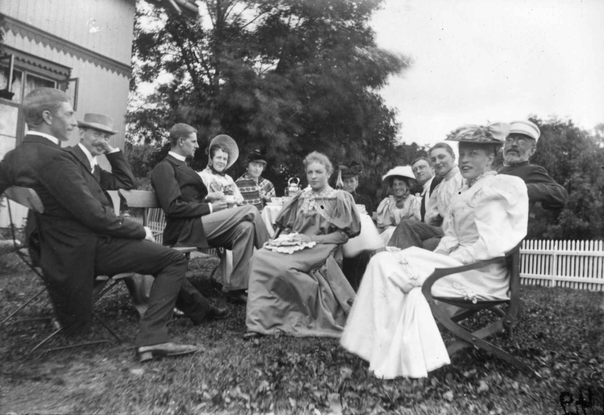 Hermann Christian Neupert, Div. drakt, Norge 1908-10. Kvinner og menn sittende i en hage. Mennene i dress. Kvinnene i lange kjoler. Noen med hatter og luer.