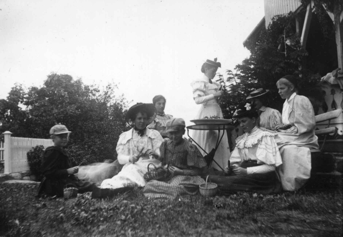 Hermann Christian Neupert, Div. drakt, Norge 1908-10. I en hage. Kvinner og barn rundt et bord. Kvinnene med hatter og i lange kjoler eller skjørt og bluse. Noen med hatter. To barn med luer i forgrunnen.