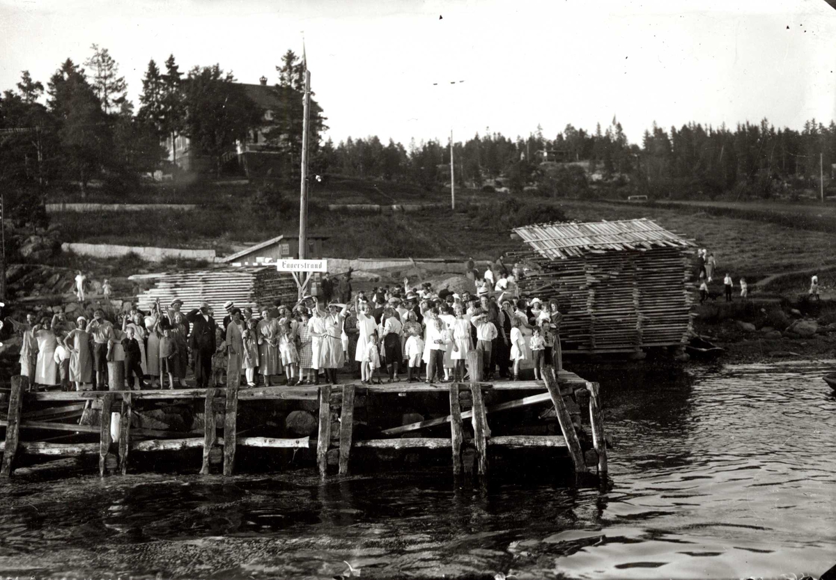 Hermann Christian Neupert, Fagerstrand, Nesodden, Akershus. 1924. Oversiktsbilde. Fergekai med folkemengde. Barn, kvinner og menn. Bebyggelse i bakgrunnen.