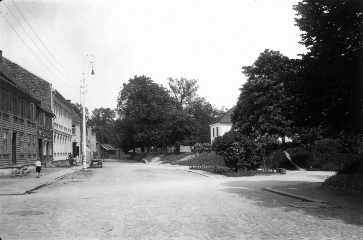 Hermann Christian Neupert, Fredrikstad, Østfold. 1930. Gate i Gamlebyen. Grøntareal til høyre. Gate med en husrekke til venstre. I bakgrunnen fotgjengere.