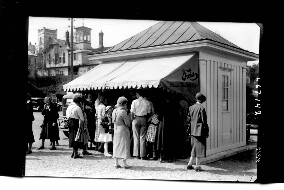 Hermann Christian Neupert, Kvinner, menn og barn foran kiosk med Freiareklame. Parkert bil. Bebyggelse i bakgrunnen.