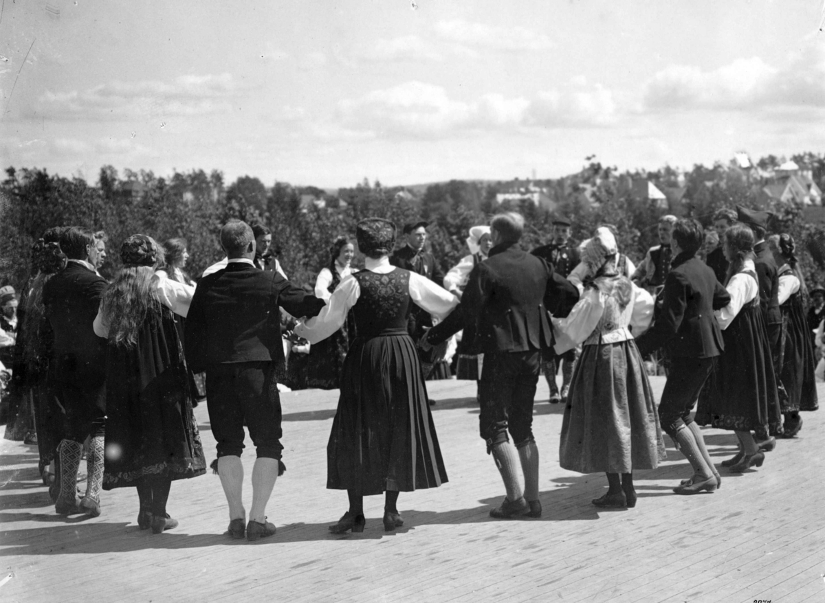 Hermann Christian Neupert, Folkedansoppvisning på en scene. Publikum. Trær og bebyggelse i bakgrunnen.