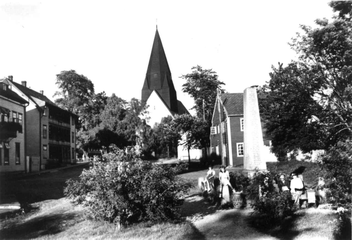 Hermann Christian Neupert, Voss kirke. 1939. Såvidt synlig bak trær. En liten park med omkringliggende hus. Kvinner og barn sittende på benker i forgrunnen..