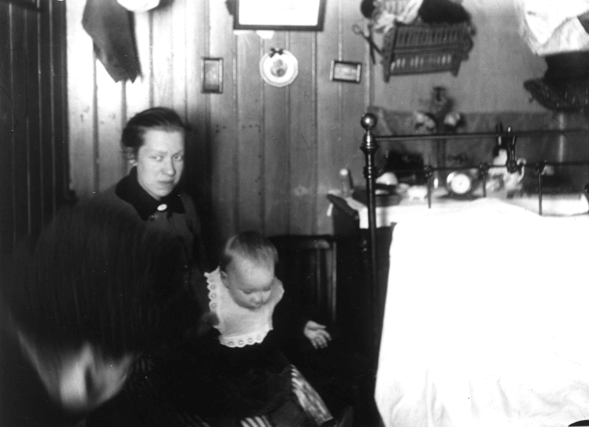 Nanna Broch, Interiør, Oslo. Kvinne og barn sitter ved seng i tett møblert rom. Fra boliginspektør Nanna Brochs boligundersøkelser i Oslo 1920-årene.