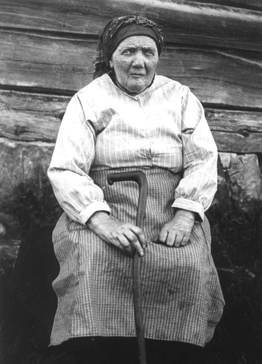 Anna Grostøl, Portrett av en ukjent kvinne i hverdagsklær og med stokk, hun sitter foran en husvegg. Ukjent sted 1936.