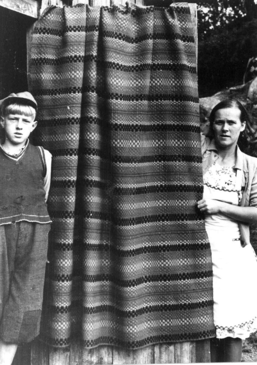 Anna Grostøl, Iveland, Vest-Agder 1941. Teppe, muligens tavlebragd, spunnet og vevd ca. 1900. To personer står ved siden av det hengende teppet.