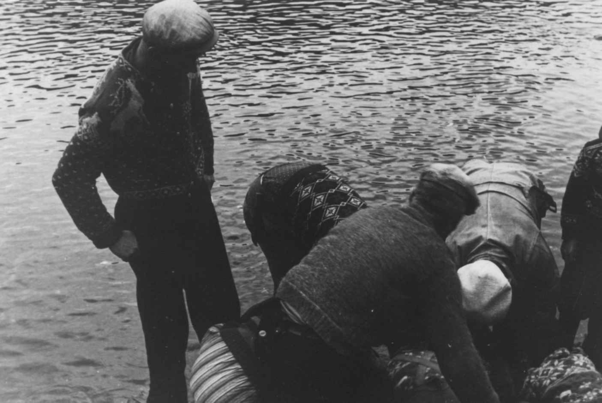 Anna Grostøl, Fiske. Nærbilde av en lite gruppe mennesker samlet om et fiskegarn i strandkanten. Åseral, Vest- Agder 1943.