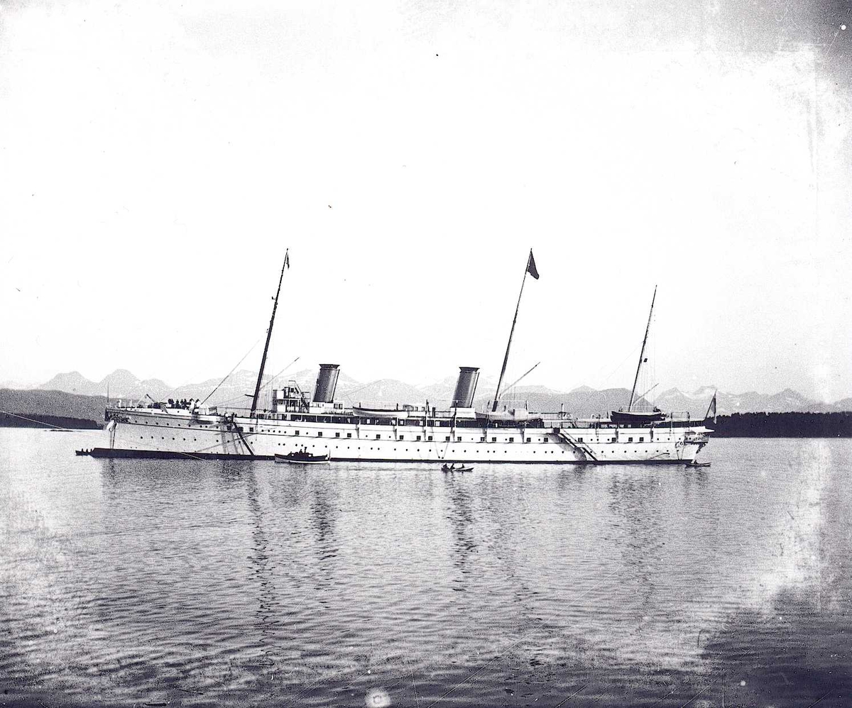 Tom Kent, Keiser Wilhelm II' skip S.M.S. Hohenzollern på havna i Molde, under et av sine årlige besøk i perioden 1889-1914. Motorbåt og robåt på siden av skipet. Sunnmørsalpene i bakgrunnen.