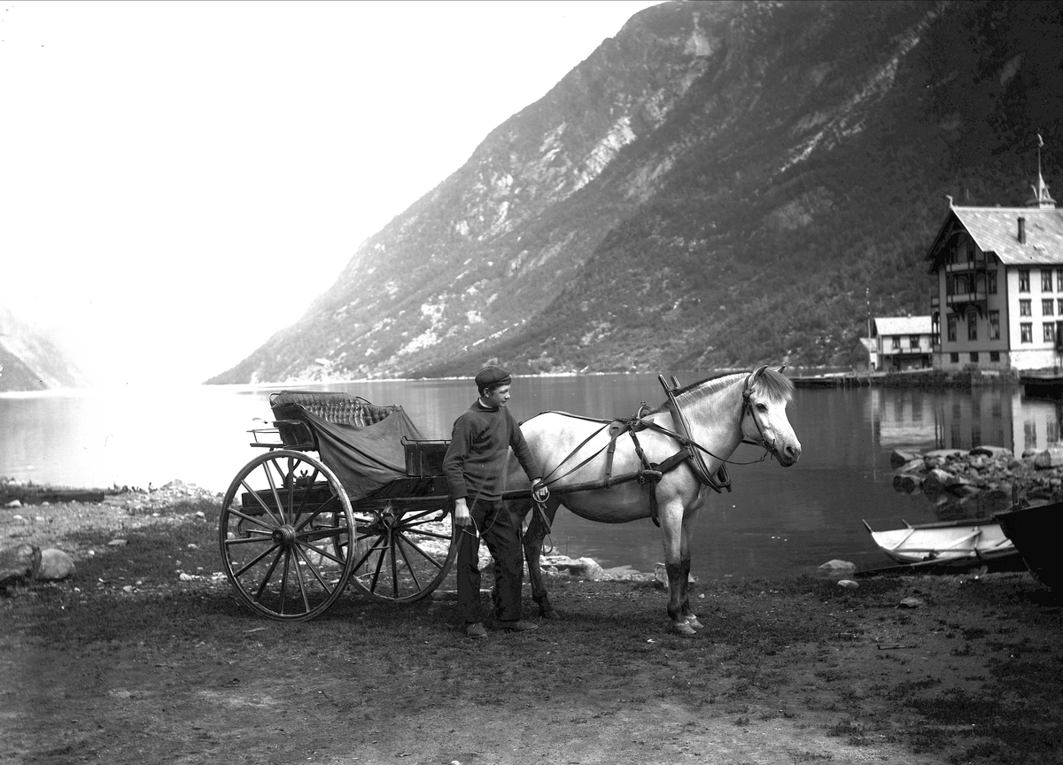 Solveig Lund, Odda i Hardanger. Gutt med hest og kjerre står foran Sørfjorden. Til høyre i bildet sees to robåter og bak disse Hardanger Hotell. Fjellandskap i bakgrunnen.