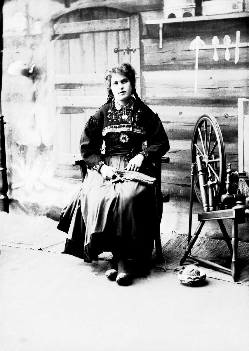 Solveig Lund, Studioportrett av kvinne i nasjonaldrakt. Kvinnen sitter og strikker, ved siden av henne står en rokk. Kvinnen ser på betrakteren. På gulvet står en bolle med et garnnøste. Bakgrunnen er malt og viser seterinteriør. Til venstre står kjelen på ildstedet og til høyre står vegghylle med tiner.