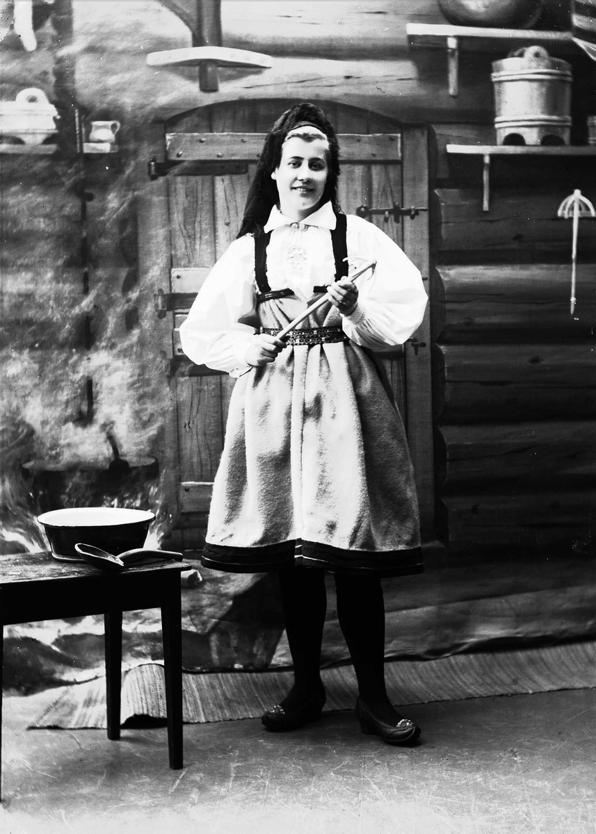 Solveig Lund, Studioportrett av kvinne i nasjonaldrakt. Hun står med en sleiv i hendene ved siden et bord med bolle og sleiv. Kvinnen ser ut mot betrakteren. Bakgrunnen er malt og viser seterinteriør. Til venstre står kjelen på ildstedet og til høyre står vegghylle med tiner.