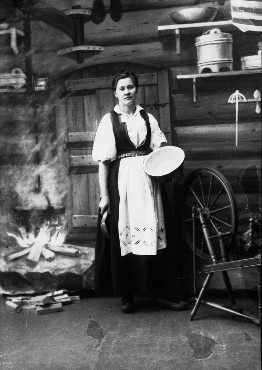 Solveig Lund, Studioportrett av kvinne i nasjonaldrakt. Hun står med en sleiv og bolle i hendene. Ved siden av henne står en rokk på gulvet. Kvinnen ser nedover. Bakgrunnen er malt og viser seterinteriør. Til venstre står kjelen på ildstedet og til høyre står vegghylle med tiner.