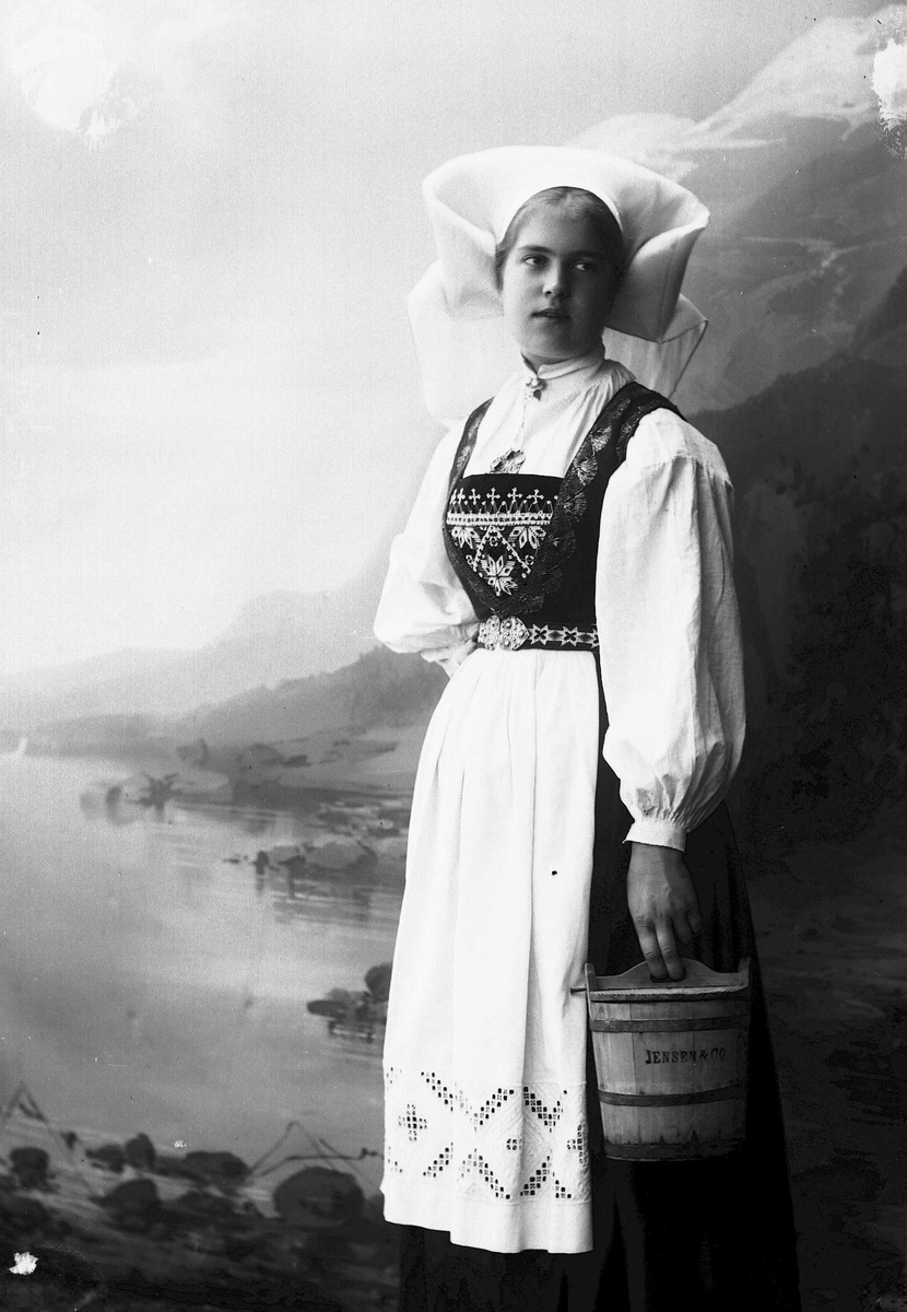 Solveig Lund, Studioportrett av kvinne i nasjonaldrakt som holder tine.Ser mot høyre i bildet. Bakgrunnen er malt med fjord og fjell landskap.