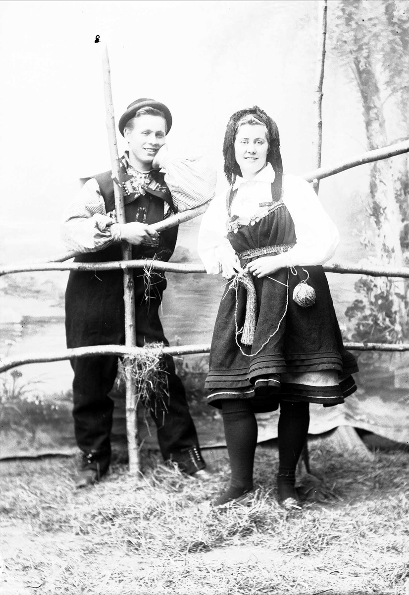 Solveig Lund, Studioportrett av kvinne og mann i nasjonaldrakter. Mannen lener seg mot hesjegjerde og ser på kvinnen som holder strikketøy i hendene. Bakgrunnen er malt med skog, vann og bladverk