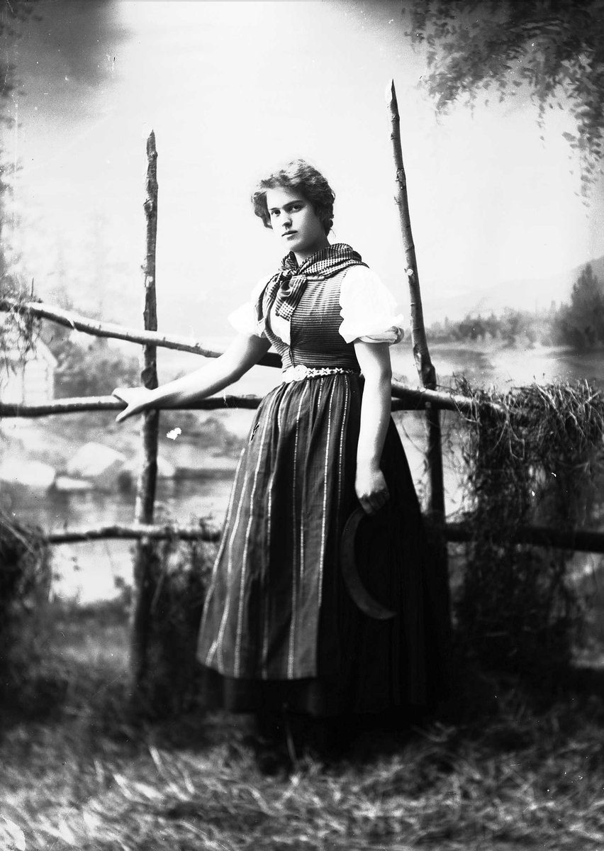 Solveig Lund, Studioportrett av kvinne i nasjonaldrakt. Hun lener seg mot hesjegjerde og holder sigd i hånden. Bakgrunnen er malt med skog, vann og hytte. På gulvet er det høy/gress.