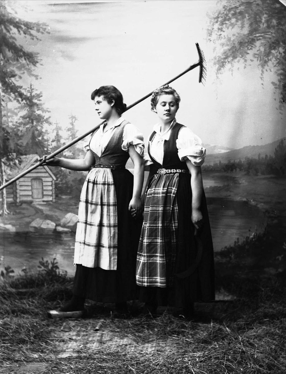 Solveig Lund, Studioportrett av to kvinner i nasjonaldrakter som holder hverandre i hendene.Den ene holder en rive over skulderen. Bakgrunnen er malt med skog, vann og hytte. På gulvet er det høy/gress.