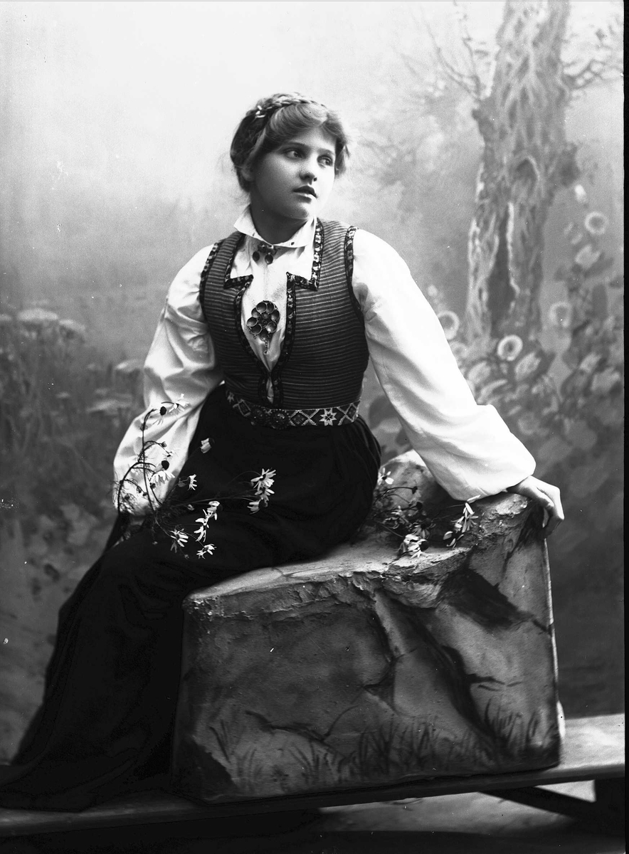 Solveig Lund, Studioportrett av kvinne i drakt sittende på modell av stein. Hun holder blomster i hendene.Bakgrunnen er malt med trær og blomster.