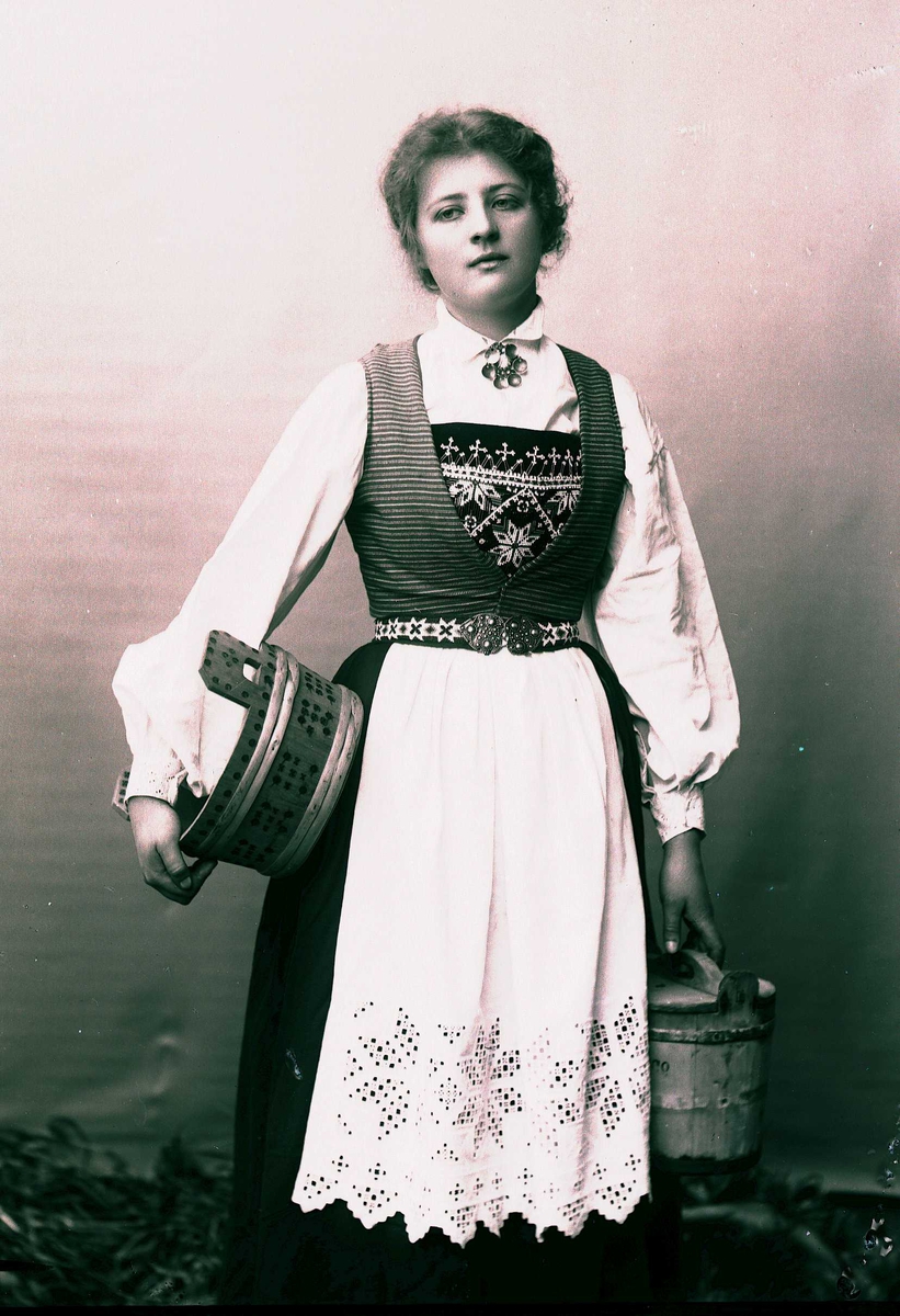 Solveig Lund, 3/4 portrett av kvinne i drakt, tatt i fotografens studio. Hun holder en tine i den venstre hånden og en under den høyre armen. Hun står på gress mot en nøytral bakgrunn.