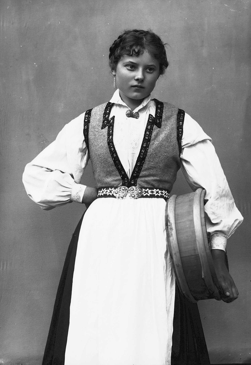 Solveig Lund, 3/4 portrett av kvinne i drakt, tatt i fotografens studio. Hun holder en melkekolle under den venstre armen og den høyre hånden på hoften. Nøytral bakgrunn.